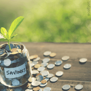 A small glass jar labelled 'SAVINGS' filled with coins, with more coins scattered around it on a wooden table. A green plant sprout is growing from the jar, symbolising financial growth and savings.