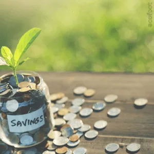 A small glass jar labelled 'SAVINGS' filled with coins, with more coins scattered around it on a wooden table. A green plant sprout is growing from the jar, symbolising financial growth and savings.