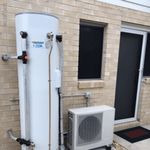An outdoor heat pump hot water system installed against a brick wall, featuring a tall water storage tank and a white heat pump unit below.