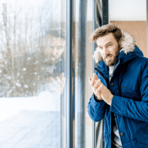 Man in a blue winter coat looking outside through a window, appearing cold and rubbing his hands for warmth during winter.