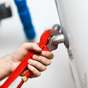 Close-up of a plumber’s hand using a red pipe wrench to tighten or loosen a fitting on a water heater unit.