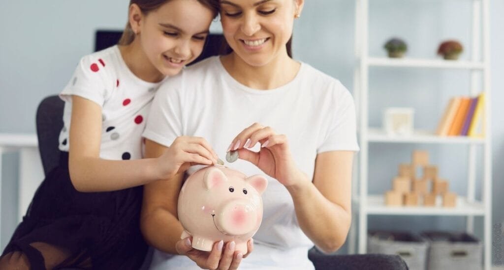 Smiling mother and daughter placing a coin into a piggy bank, symbolising savings, family, and cost benefits of heat pump water heaters.