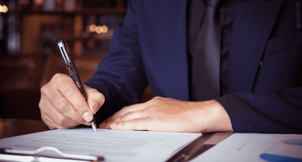 Close-up of a person writing in a notebook with a pen, sitting at a desk, wearing a dark jacket.