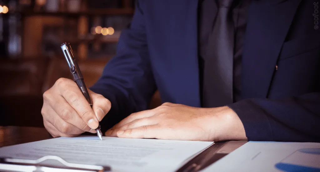 Close-up of a person writing in a notebook with a pen, sitting at a desk, wearing a dark jacket.