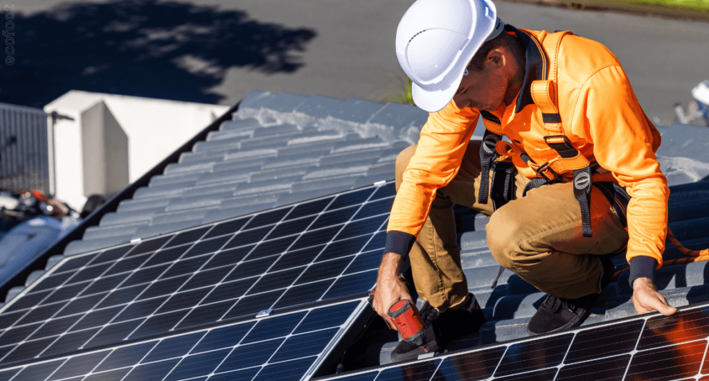 Solar installer in safety gear fitting solar panels on a tiled rooftop, using tools to secure the panels in place under bright daylight. 